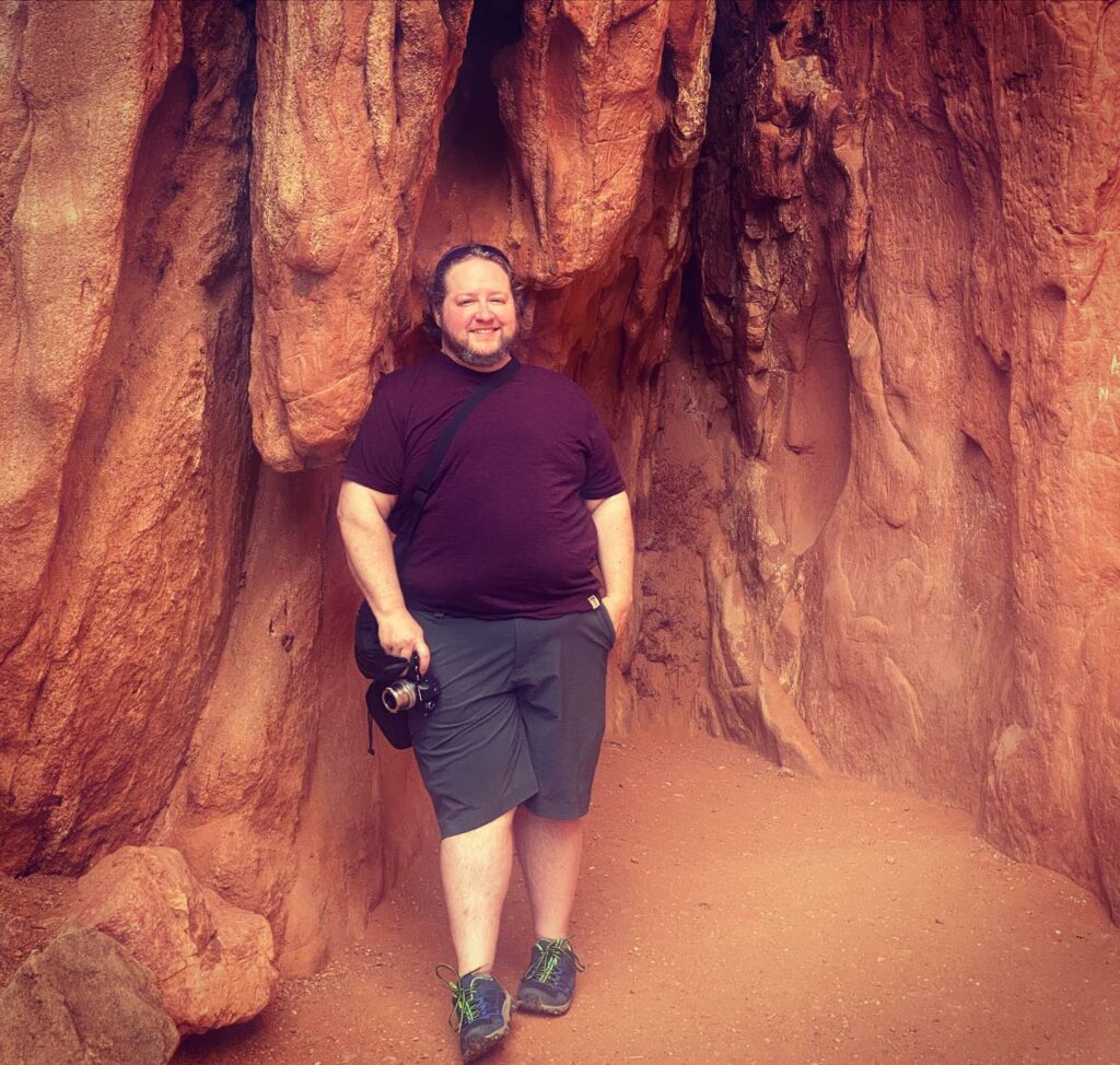 short white man in burgundy tshirt and grey shorts holding a camera and smiling, with a reddish rock formation behind him